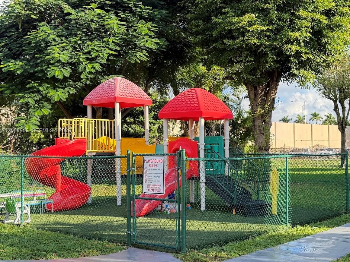 Fenced recreation area featuring a multi-level playground structure with integrated slides, stairs, and overhead canopies, set on a synthetic turf surface with mature tree coverage