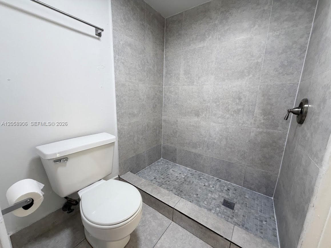 Walk-in shower featuring large-format grey wall tile, textured stone-style floor tile, and a metallic single-handle faucet, accompanied by a white porcelain toilet and a stainless steel wall-mounted fixture