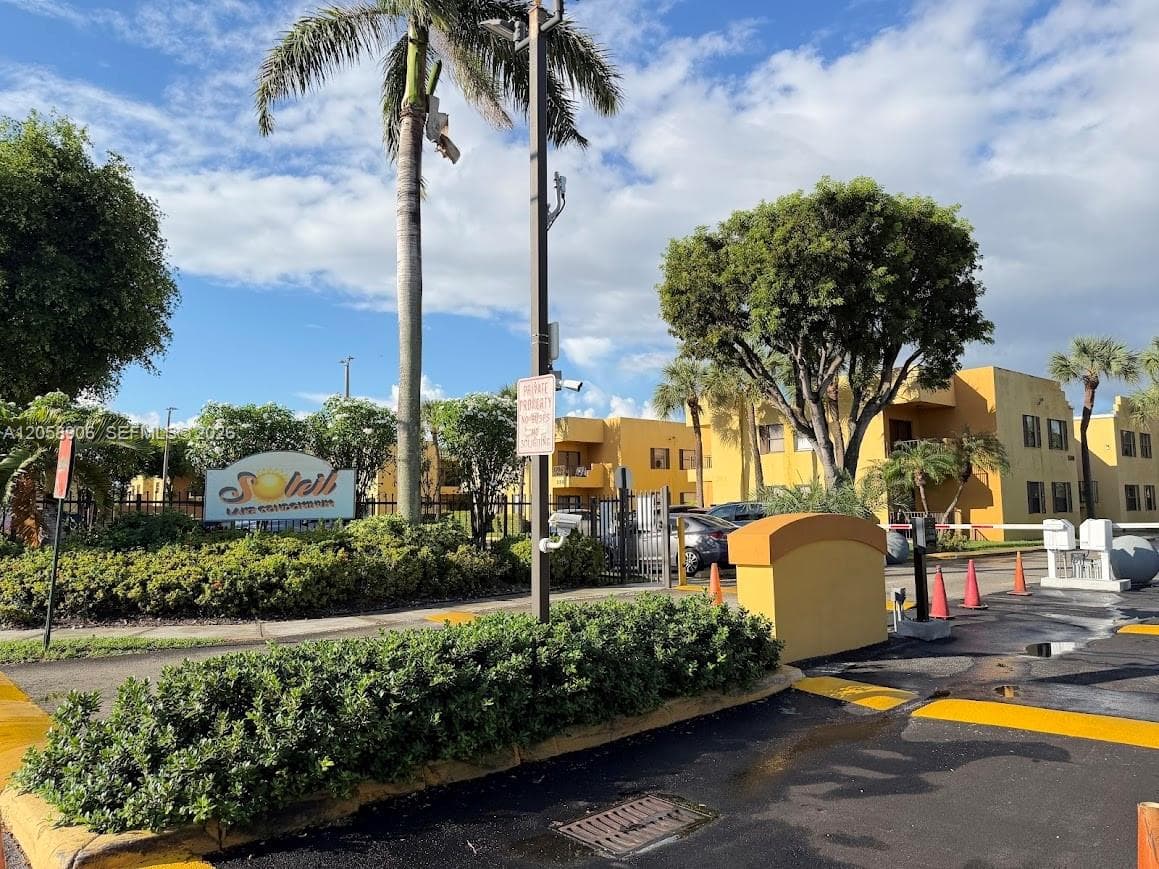 Gated entrance featuring a security access gate, palm tree landscaping, and perimeter fencing
