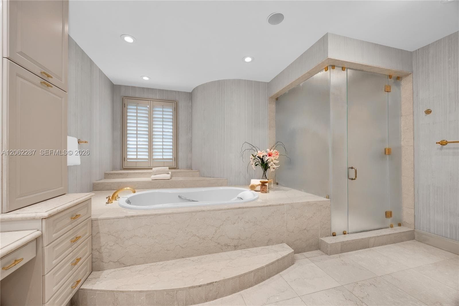 Raised stone-surround soaking tub with gold-tone fixtures, walk-in glass-enclosed shower, custom built-in cabinetry, floor-to-ceiling stone wall cladding, and recessed lighting