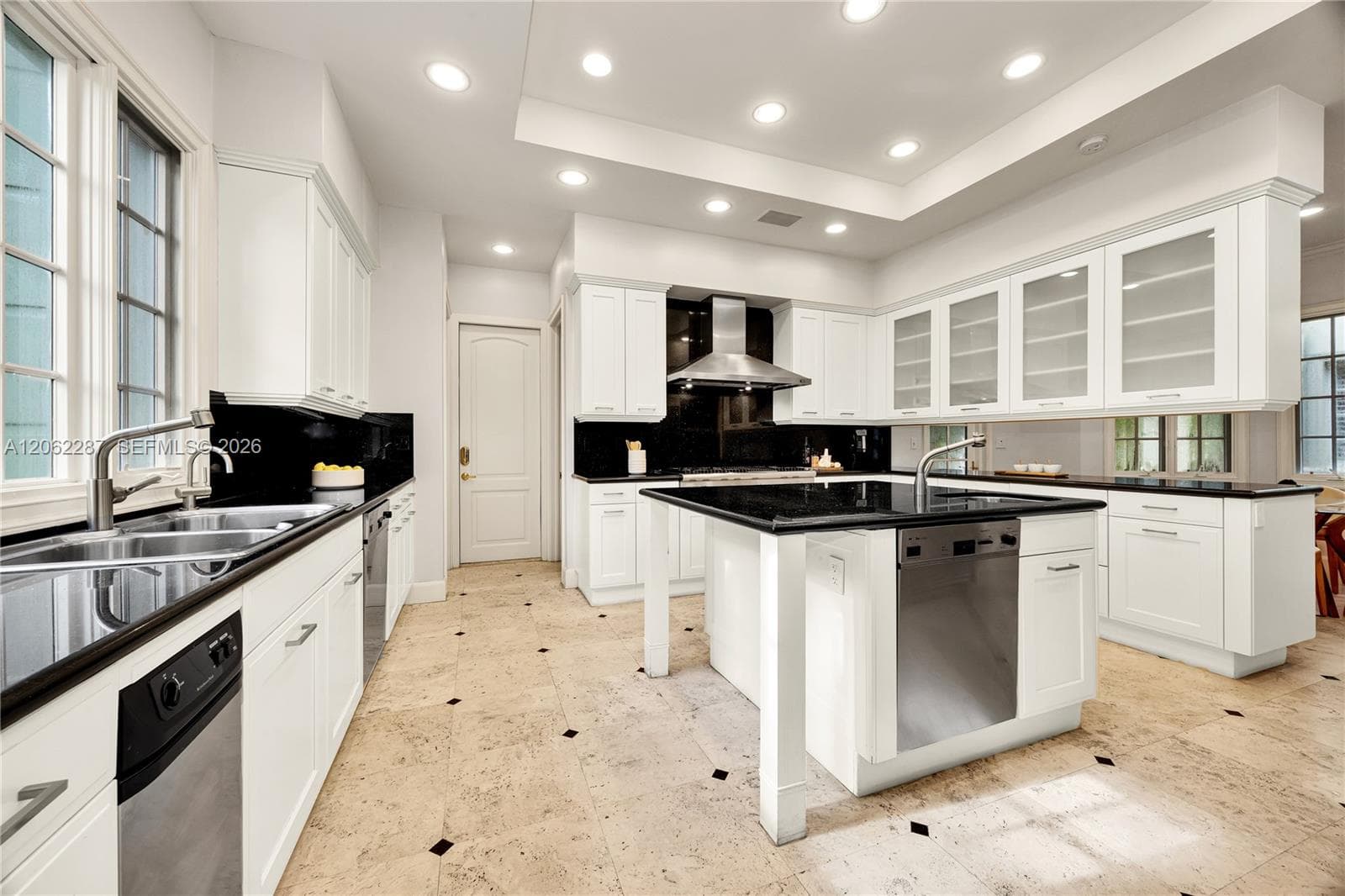 Kitchen featuring white shaker-style cabinetry, contrasting black countertops, a central island with integrated stainless steel dishwasher, recessed ceiling lighting, and stone flooring with inlaid accents
