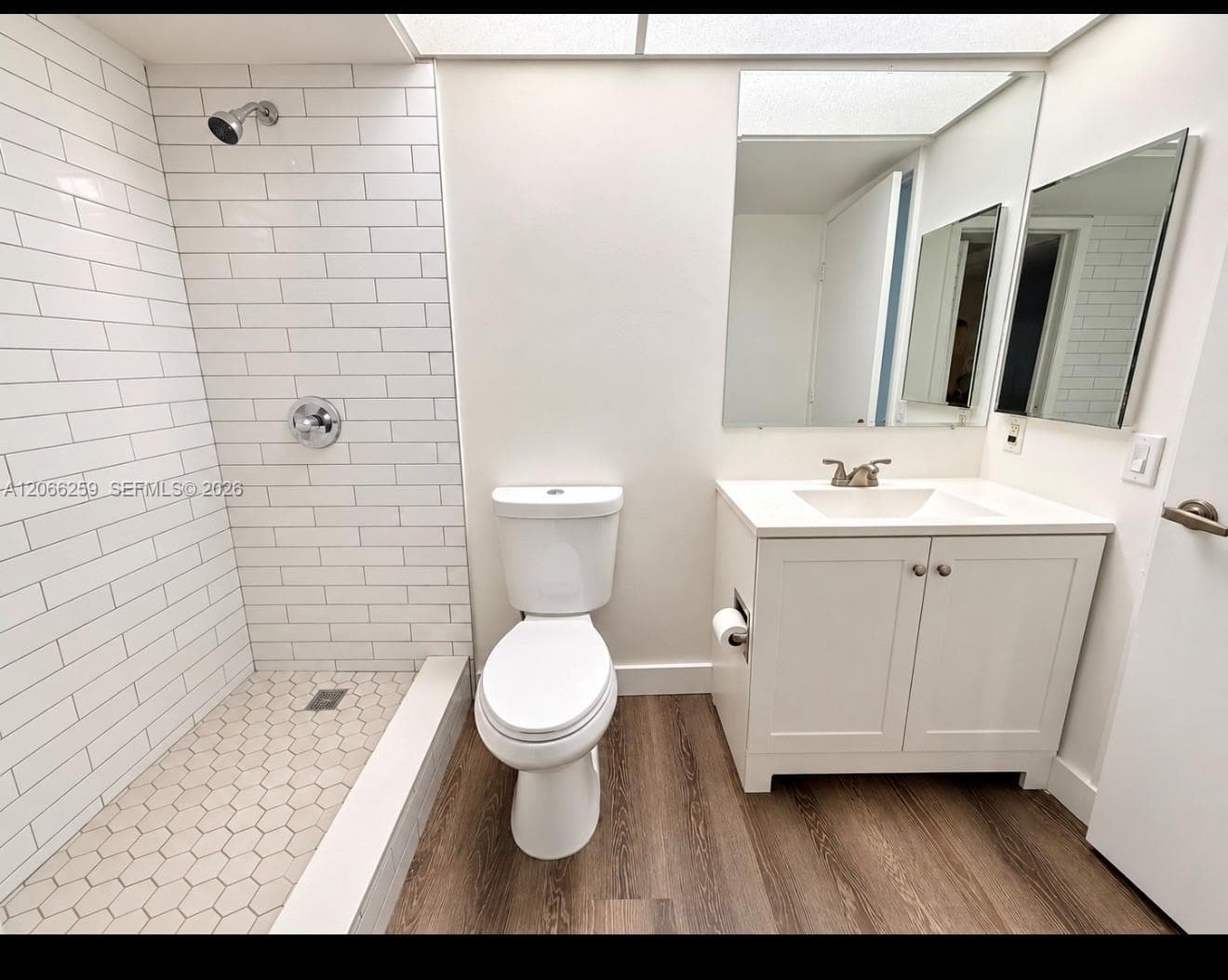 Bathroom featuring white subway tile shower surround, hexagonal floor tile, wood-finish flooring, white vanity cabinet with a integrated sink, and wall-mounted mirror