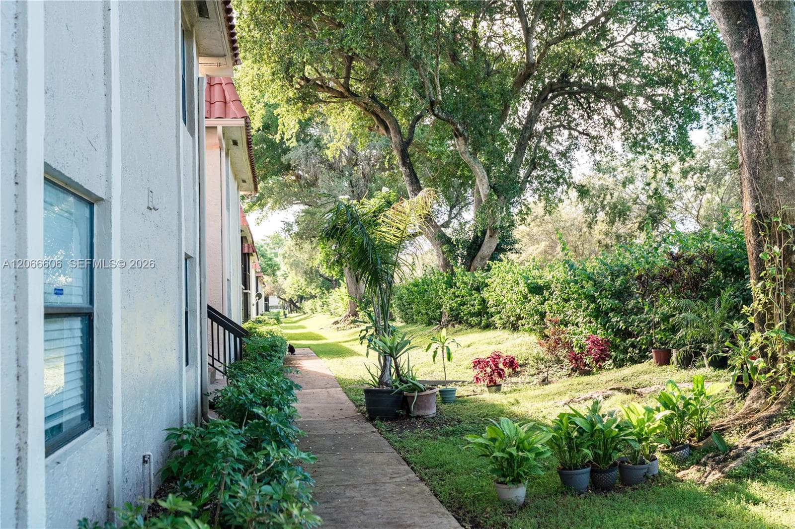 Exterior building elevation featuring textured stucco siding, red tile roofing, and a concrete pathway bordered by mature trees and landscaped greenery
