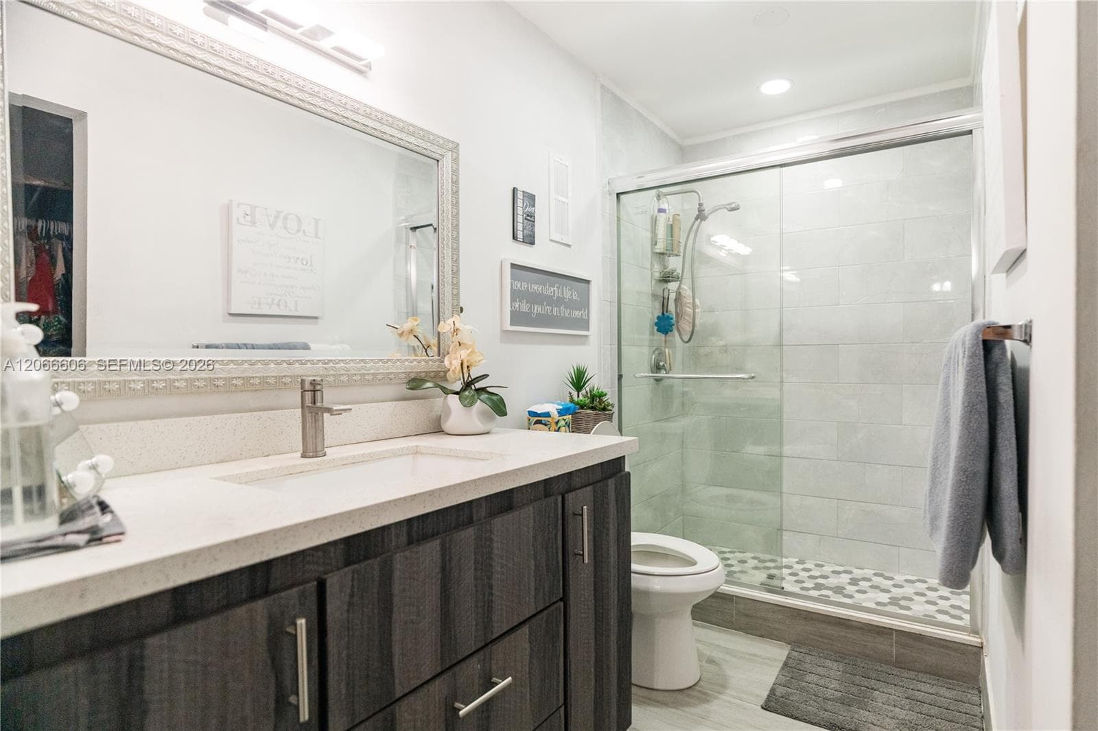 Dark wood-finish vanity with a light-toned countertop, integrated basin, large wall-mounted mirror, and walk-in shower with light tile surround and hexagonal floor tiling
