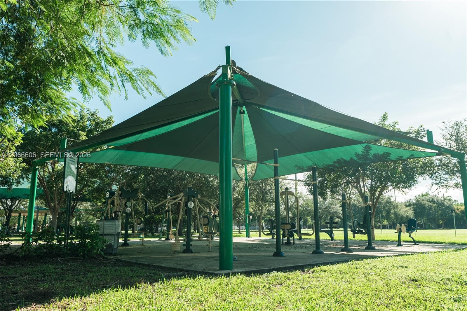 Outdoor fitness area featuring metal exercise equipment, a structural sun shade canopy, and a concrete base situated within a landscaped green space