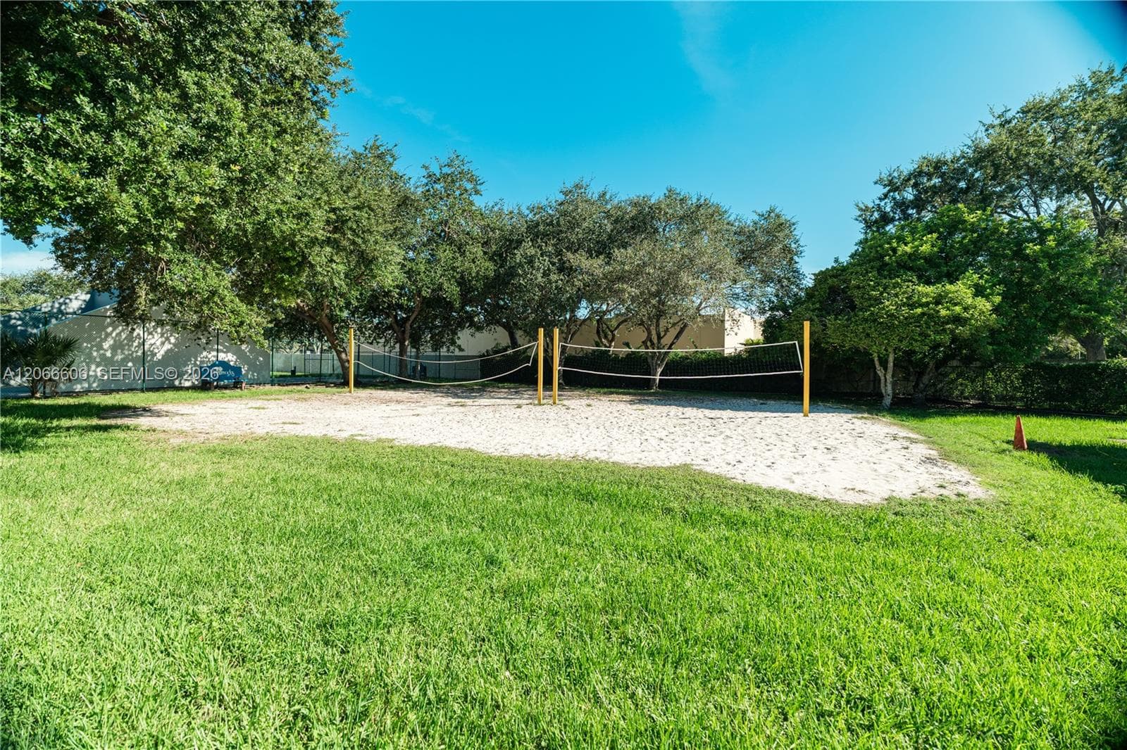 Sand volleyball court featuring a net and perimeter grass landscaping with mature trees