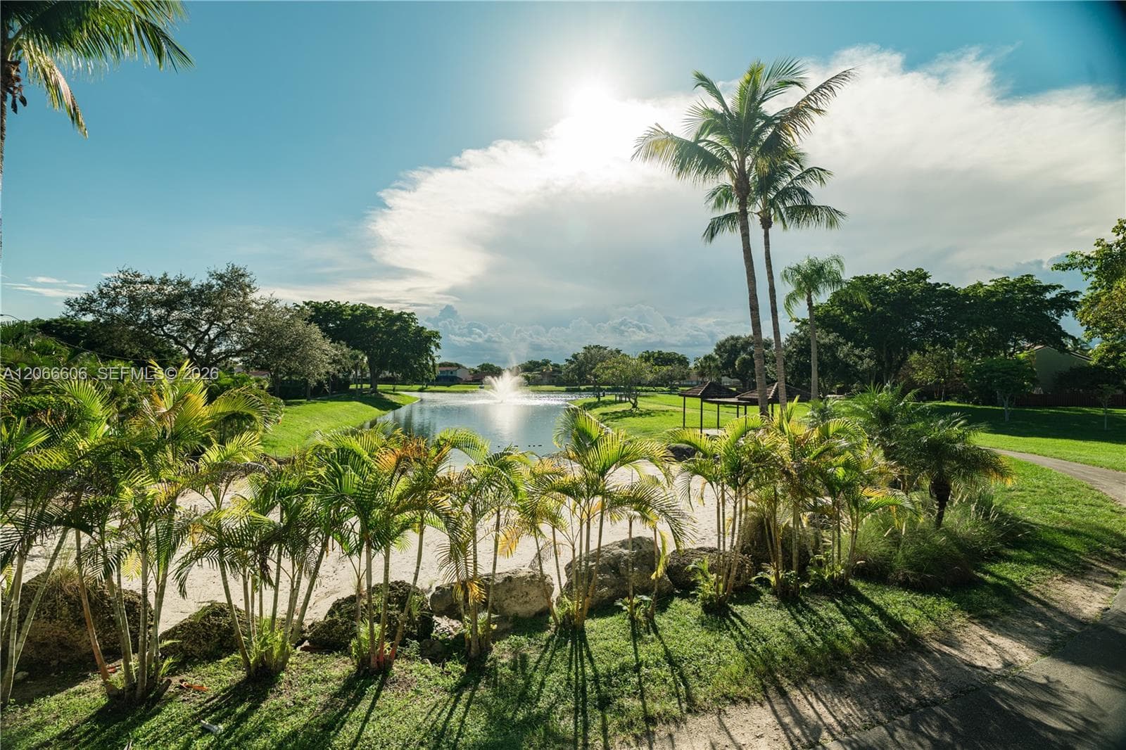 Waterfront setting with a central fountain, sandy shore, mature palm trees, rock landscaping, and paved walking paths