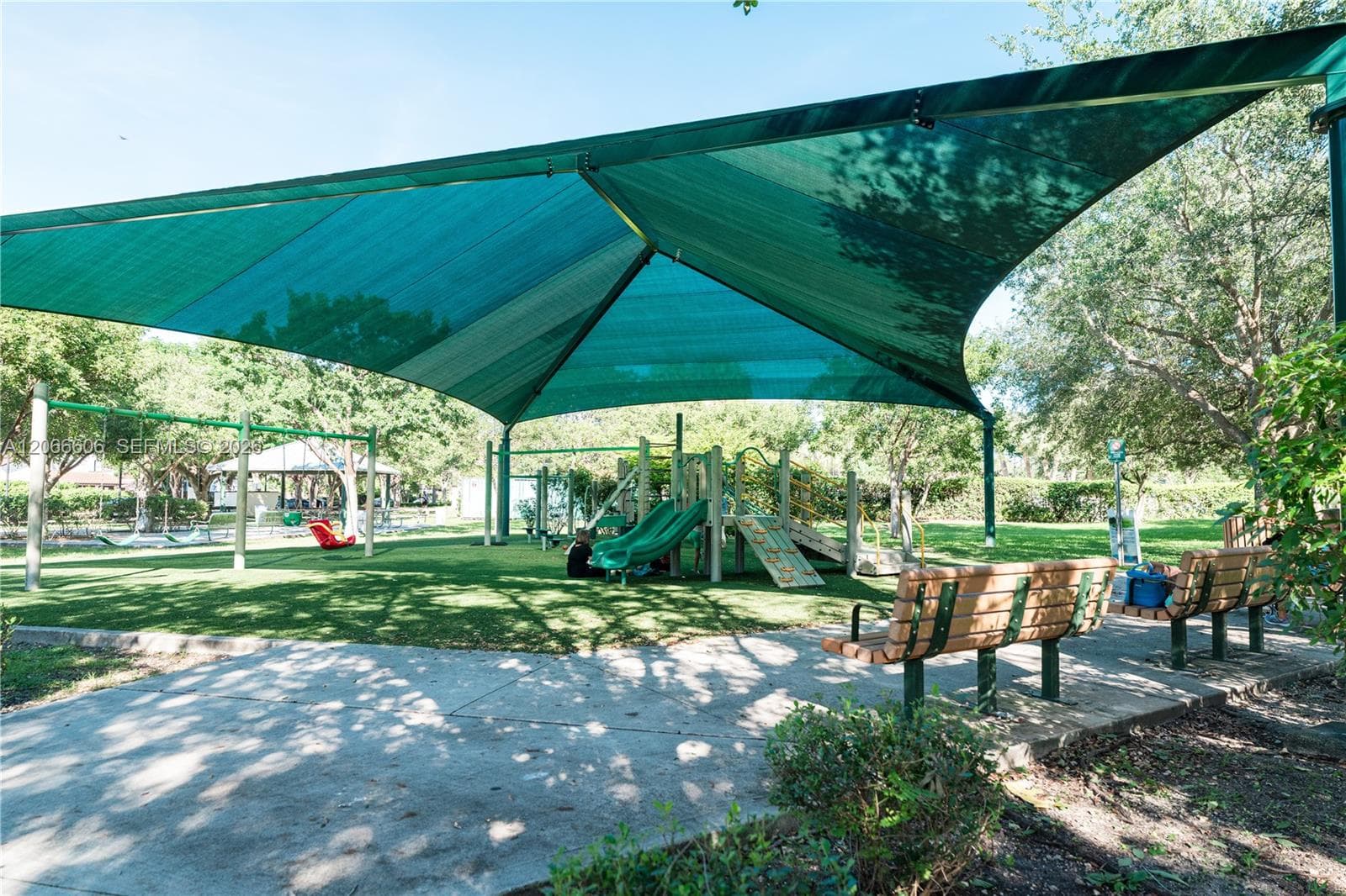 Outdoor recreation area featuring a large overhead sunshade, playground equipment, concrete pathways, and built-in wood-slat benches