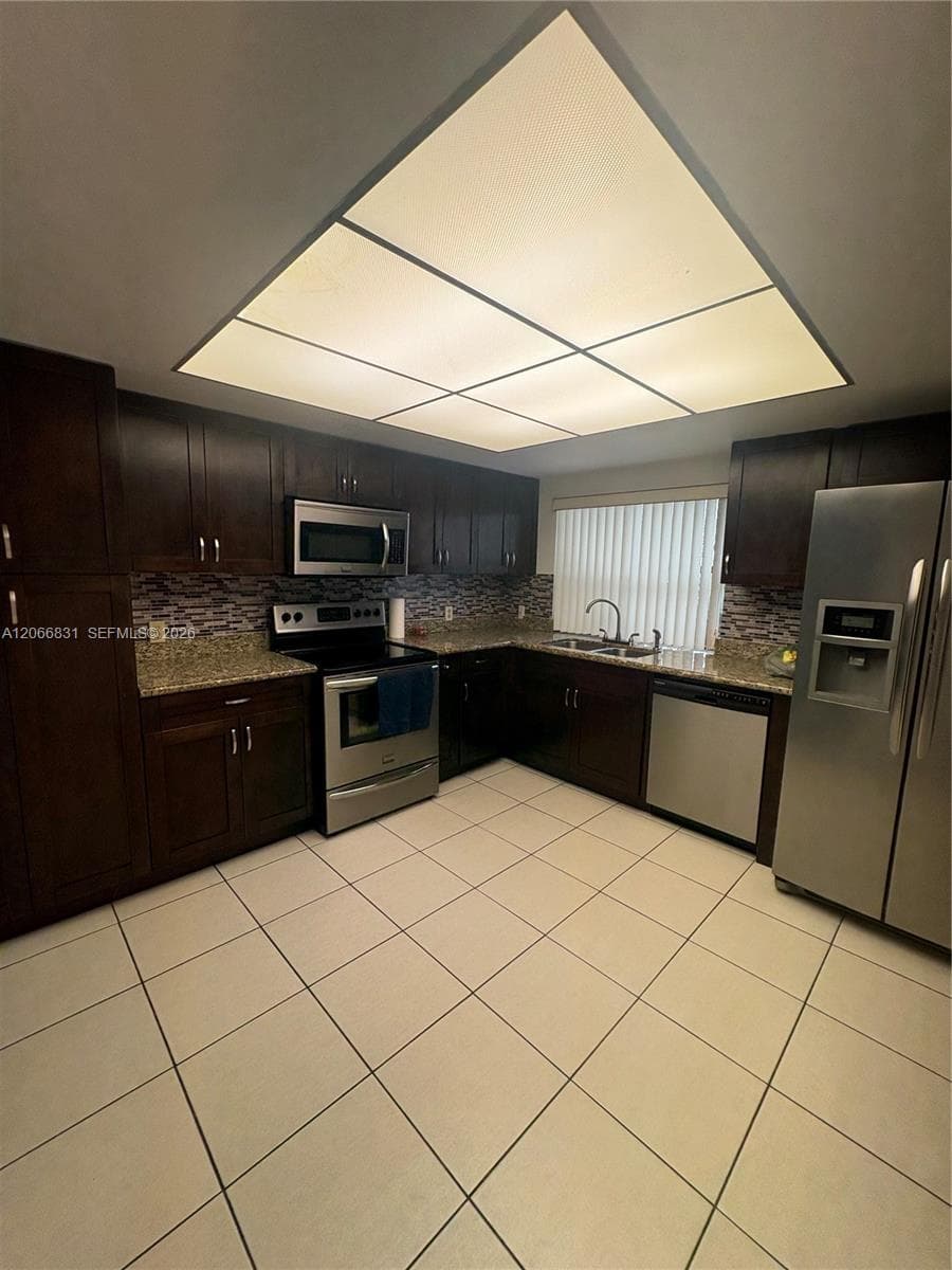 Kitchen featuring dark wood cabinetry, stone-finish countertops, stainless steel appliances, tiled backsplash, and recessed panel lighting