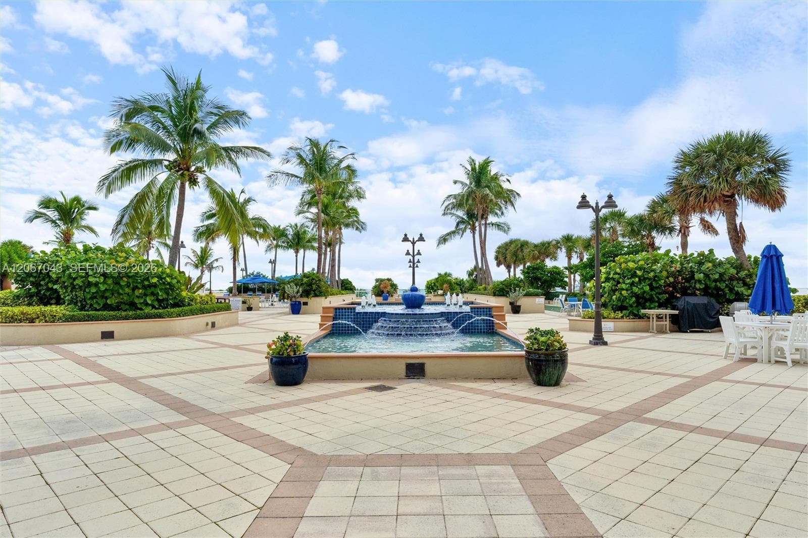 Paved plaza featuring a tiered water fountain with tiled masonry, mature palm trees, integrated landscaping, and decorative outdoor lighting