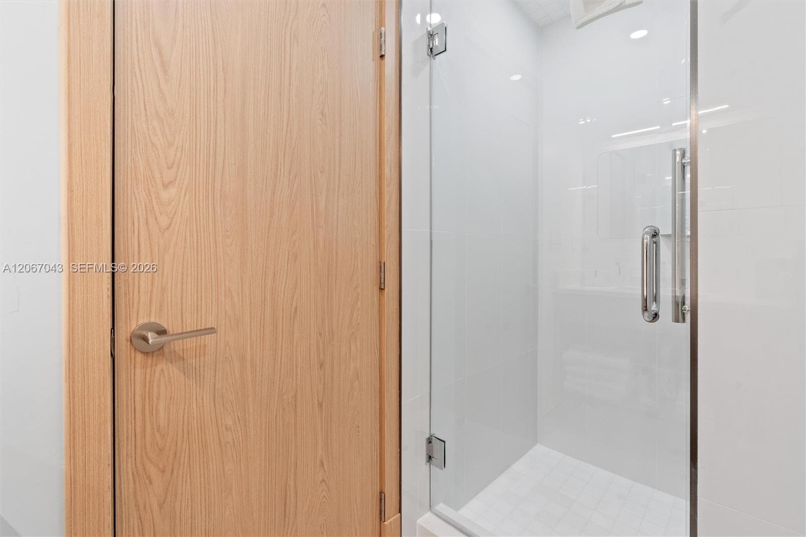 Shower enclosure with floor-to-ceiling white tile, glass door with chrome hardware, and wood-finish door with matching brushed metal handle