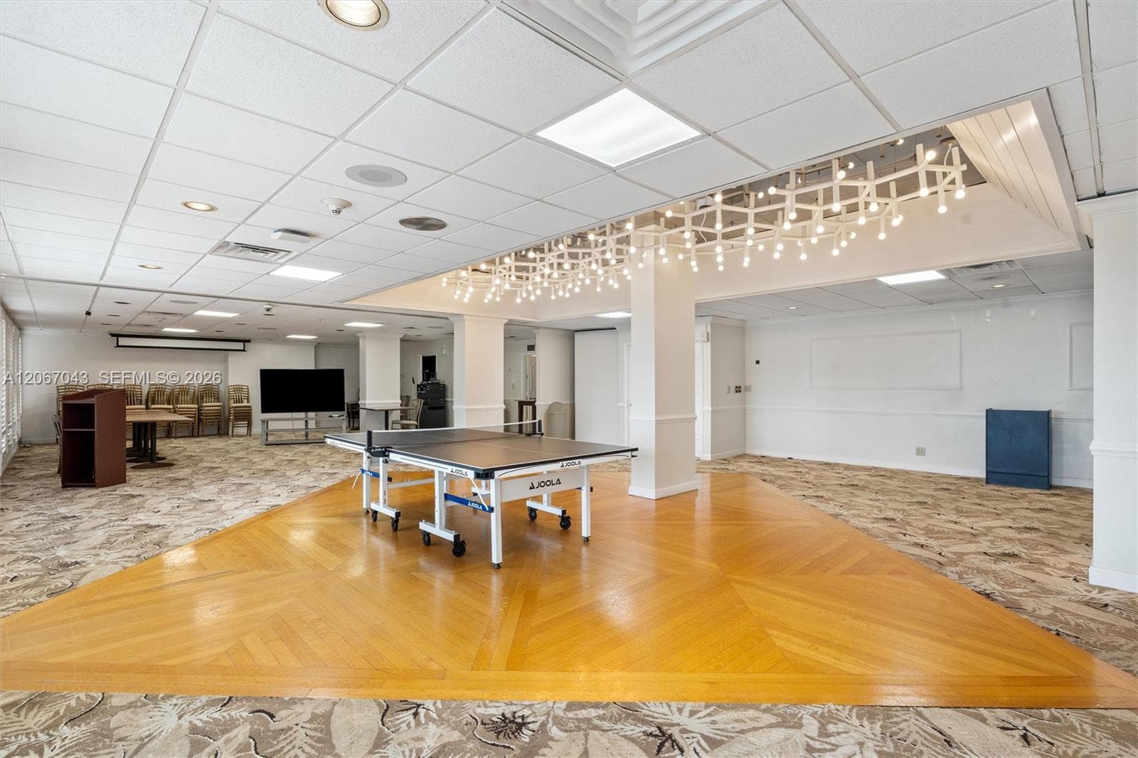 Expansive multipurpose room featuring a custom geometric chandelier, wood-finish herringbone flooring inset, patterned wall-to-wall carpeting, and recessed ceiling lighting