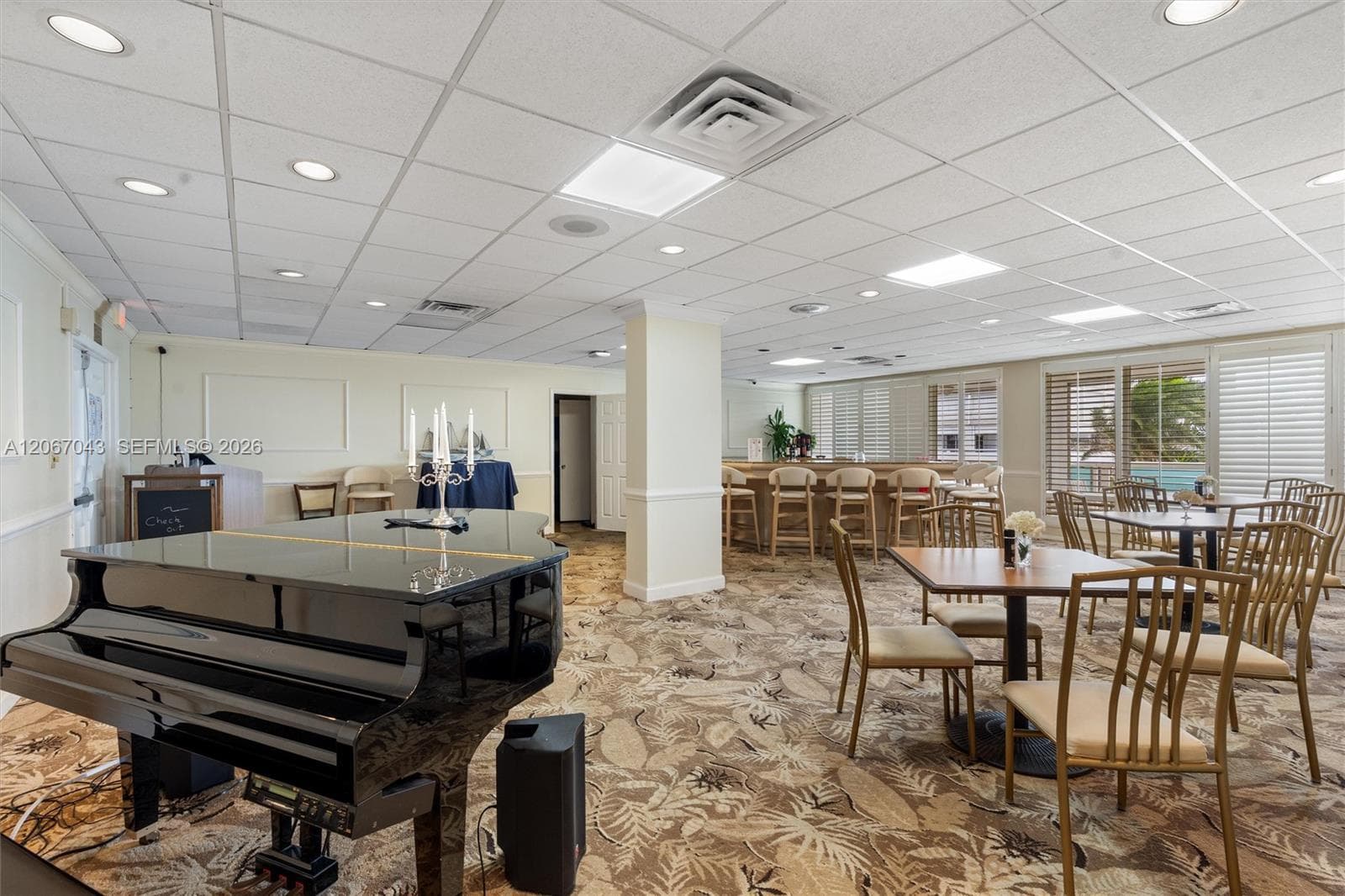 Expansive common area featuring a suspended acoustic tile ceiling, recessed lighting, integrated bar with counter seating, and floor-to-ceiling windows with horizontal blinds