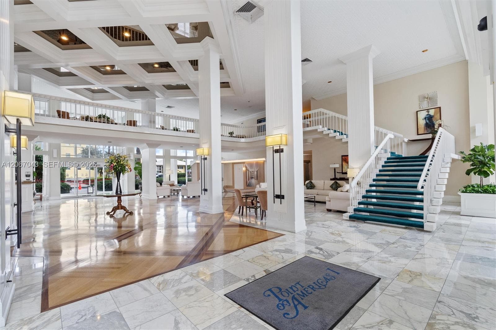 Grand atrium lobby featuring coffered ceilings, stone-tile flooring with wood inlay, structural columns, and a staircase with white railings