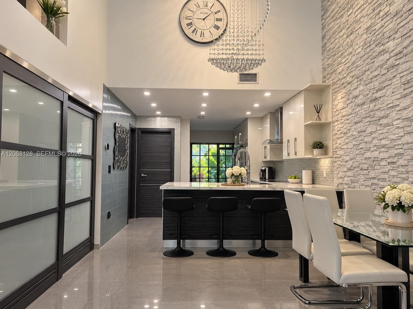 High-ceiling interior featuring a floor-to-ceiling textured stone accent wall, large-format tile flooring, a tiered crystal chandelier, and sliding doors with frosted glass panels