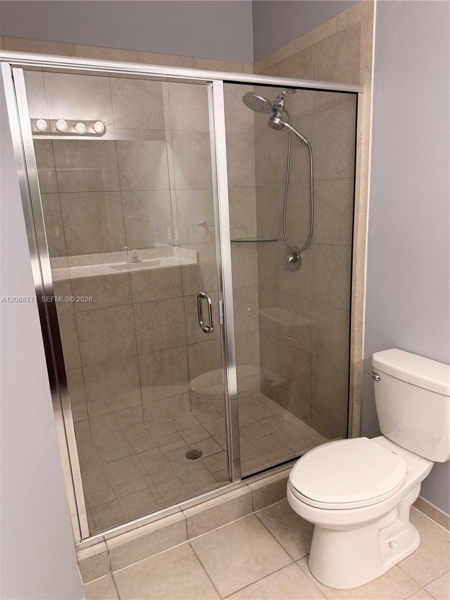 Walk-in shower featuring a sliding glass door, neutral tile surround, and adjustable handheld showerhead, accompanied by a standard-height porcelain toilet