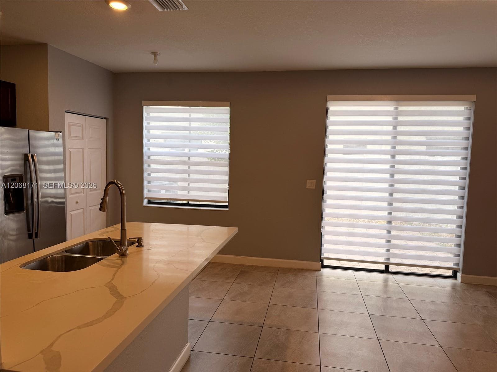 Kitchen island featuring a stone-look countertop, undermount double-basin sink, and high-arc faucet, complemented by tile flooring, stainless steel refrigerator, and dual-layered window treatments