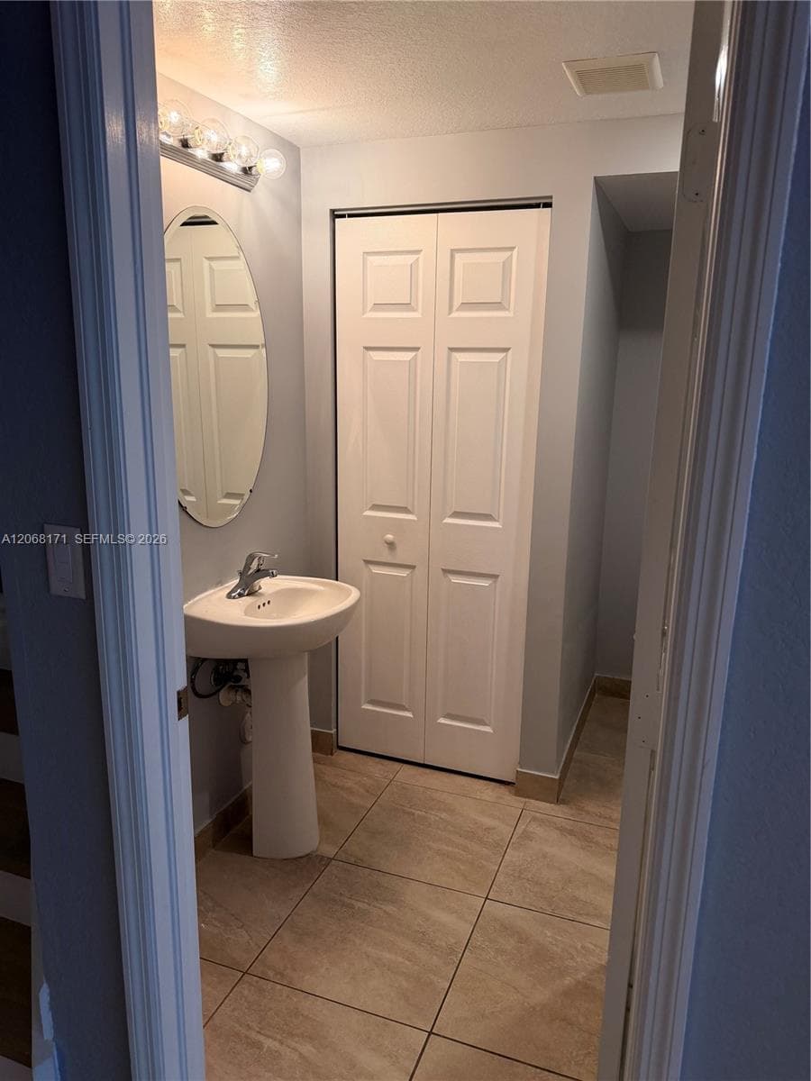 White pedestal sink with a chrome faucet, oval wall-mounted mirror, light-colored tile flooring, and bi-fold closet doors
