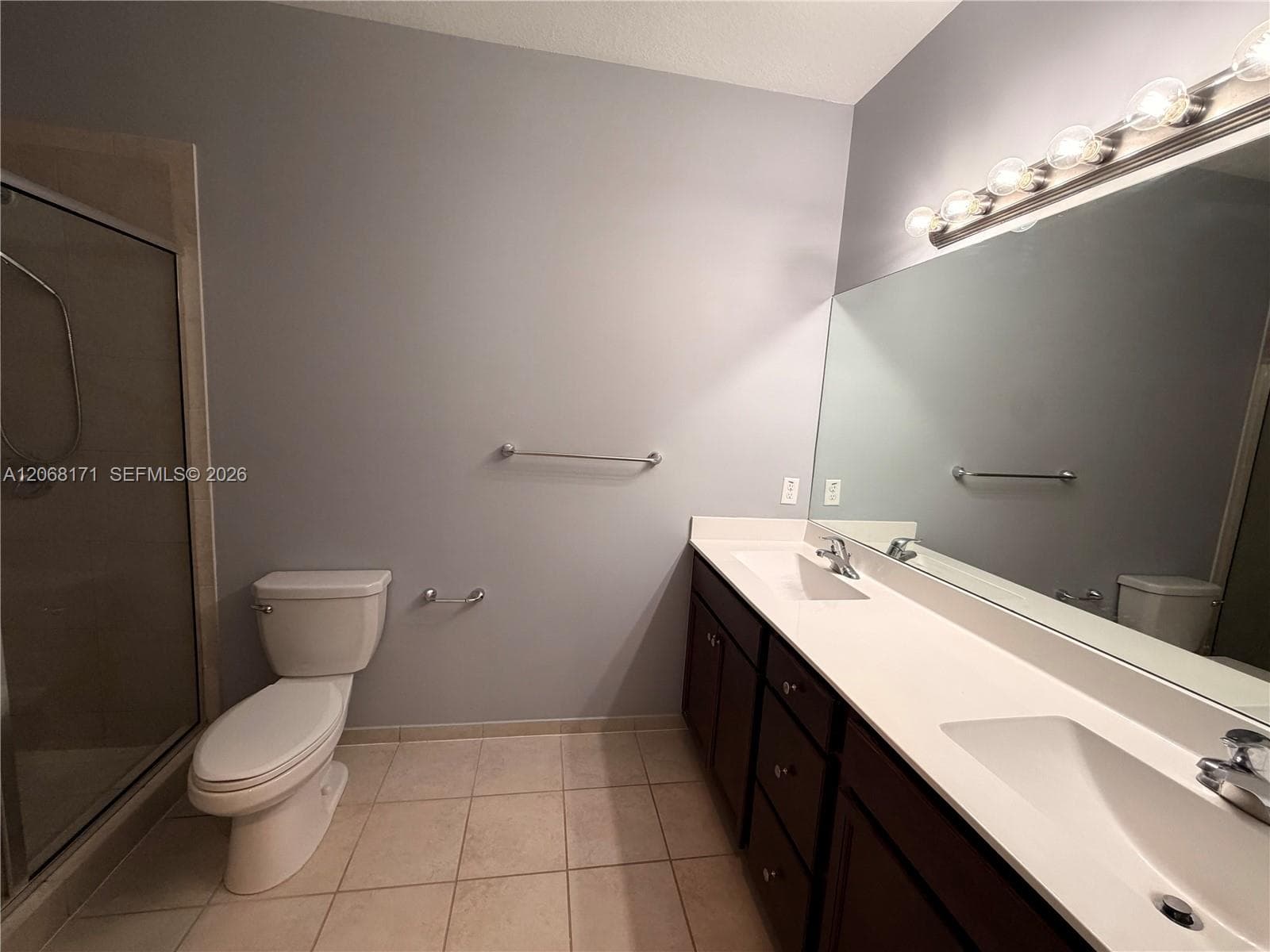Bathroom featuring a dual-sink vanity with a solid-surface countertop, dark wood-finish cabinetry, a large wall-mounted mirror with integrated light fixture, and a glass-enclosed shower