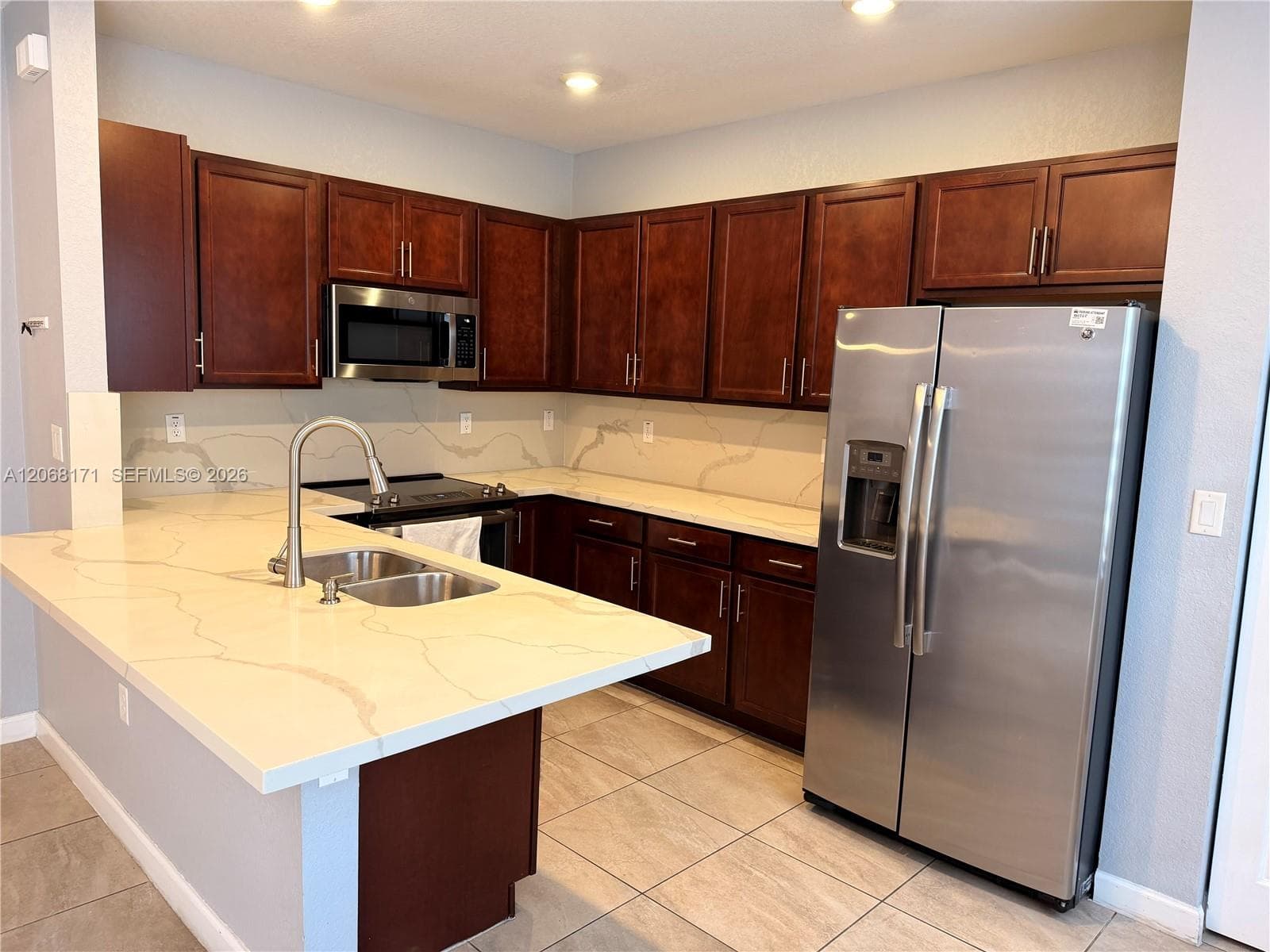 Kitchen featuring dark wood cabinetry, veined stone-finish countertops, stainless steel appliances, a peninsula with an undermount dual-basin sink, and tile flooring