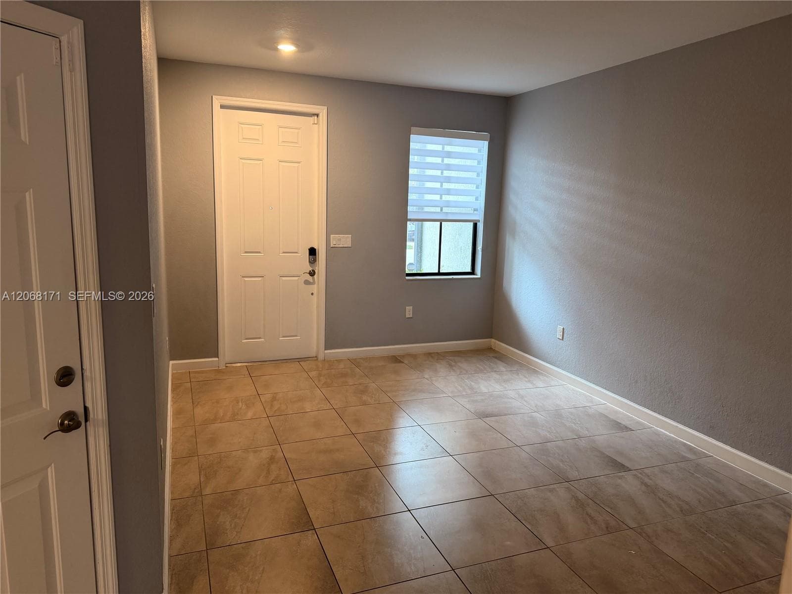 Neutral-toned interior room featuring large-format tile flooring, a six-panel entry door, and a single-hung window with a roller shade