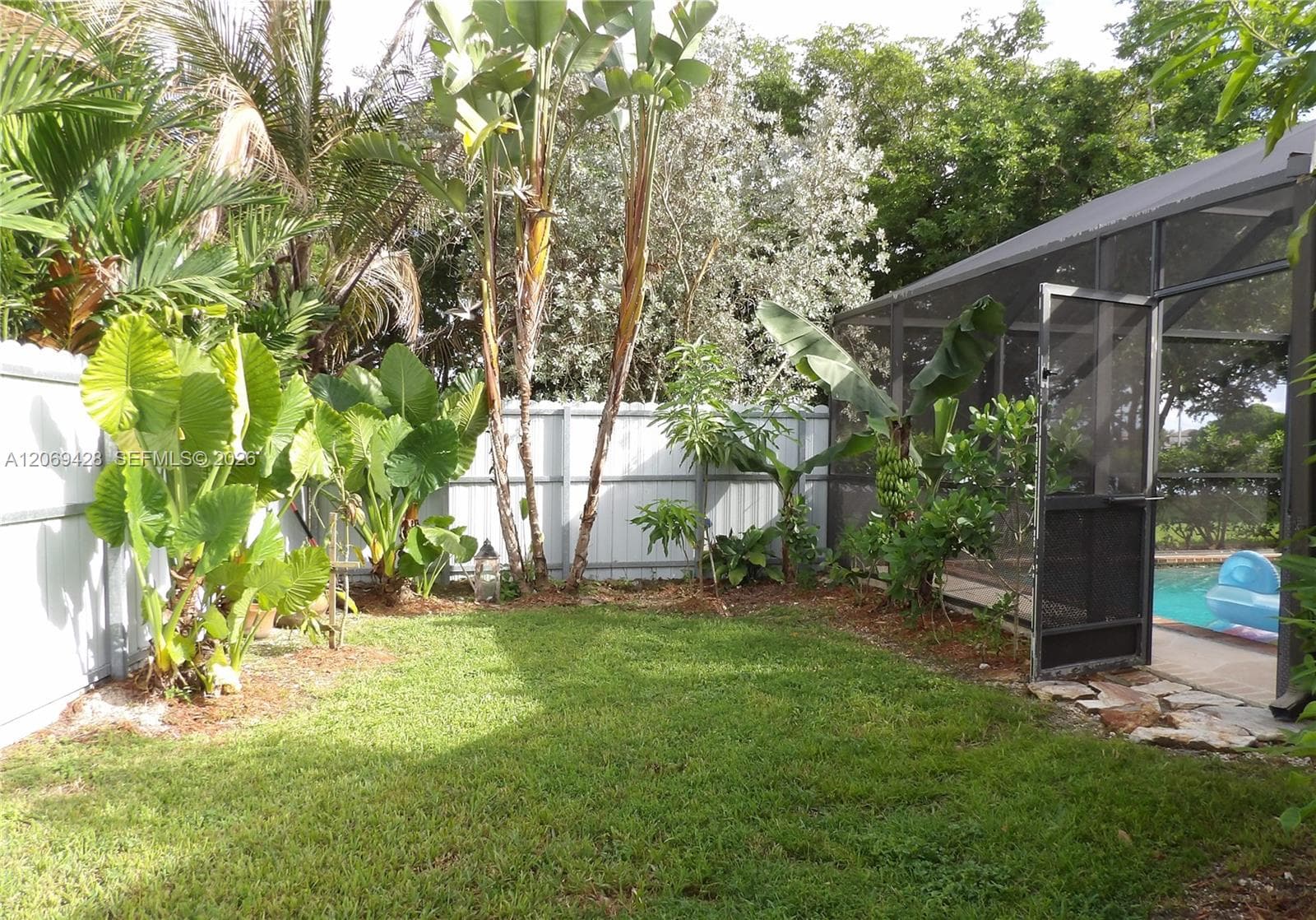 Expansive lawn area with a screened enclosure, mature tropical foliage, and a perimeter fence
