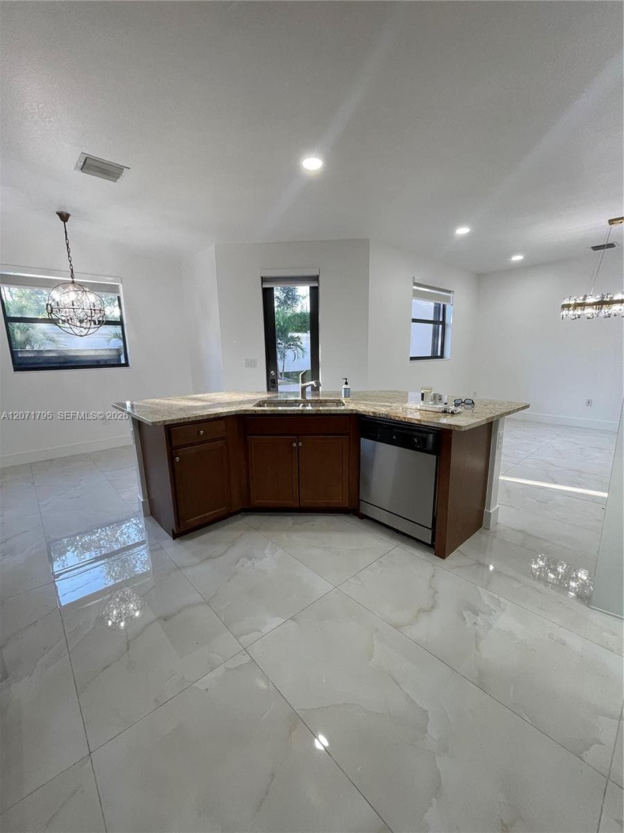 Kitchen island featuring wood cabinetry, stone-finish countertops, stainless steel undermount sink, and integrated stainless steel dishwasher, complemented by large-format tile flooring and decorative pendant lighting