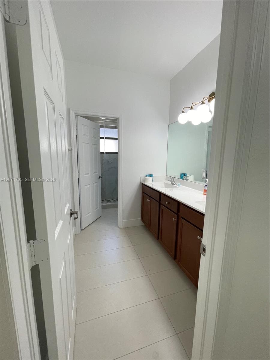 Bathroom featuring a dual-sink vanity with wood-finish cabinetry, solid surface countertop, large wall-mounted mirror, and multi-globe vanity lighting, complemented by tile flooring