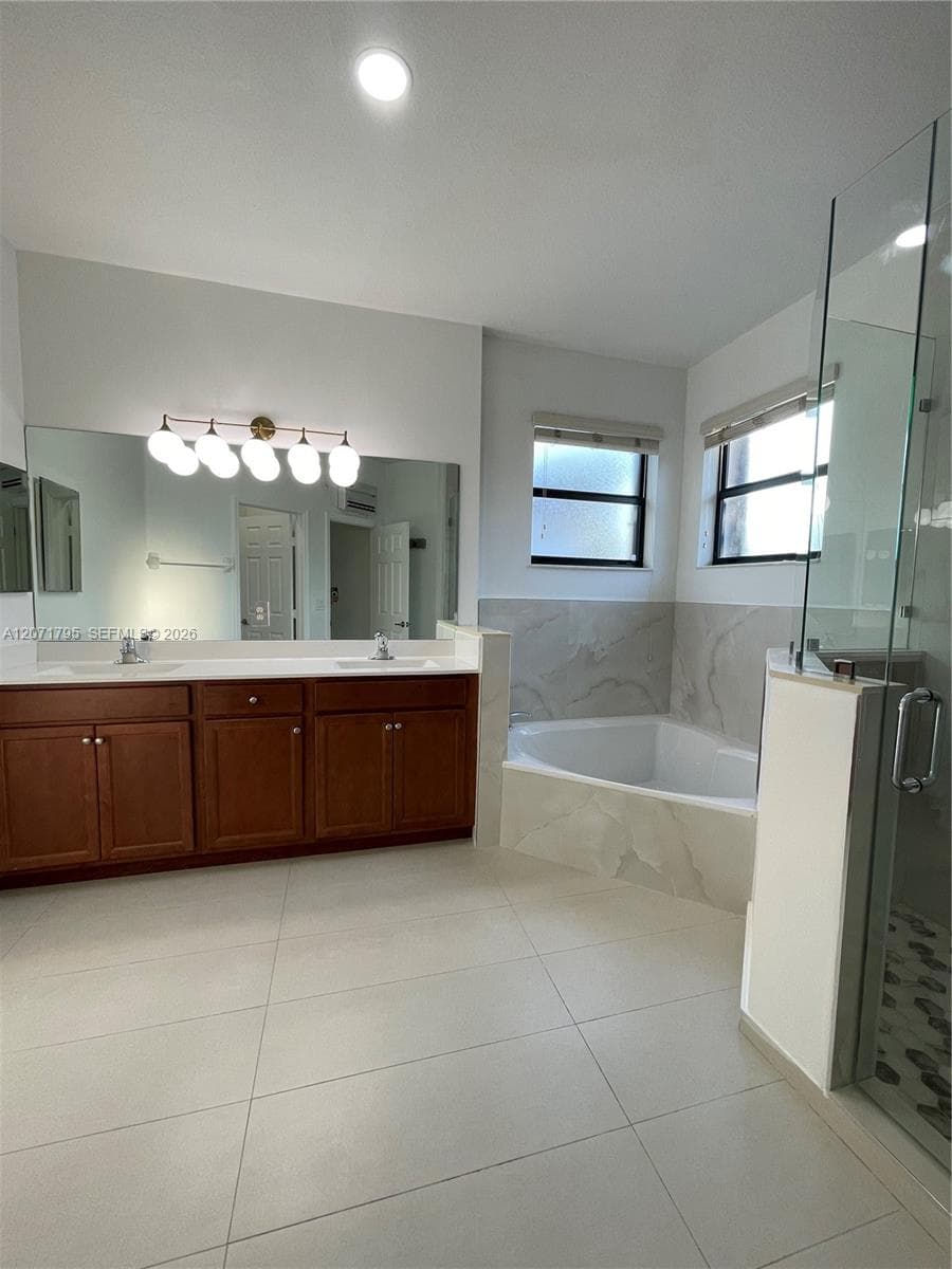 Bathroom featuring a double-sink vanity with wood cabinetry, a large wall-mounted mirror, a separate glass-enclosed shower, and a built-in soaking tub with surround tiling