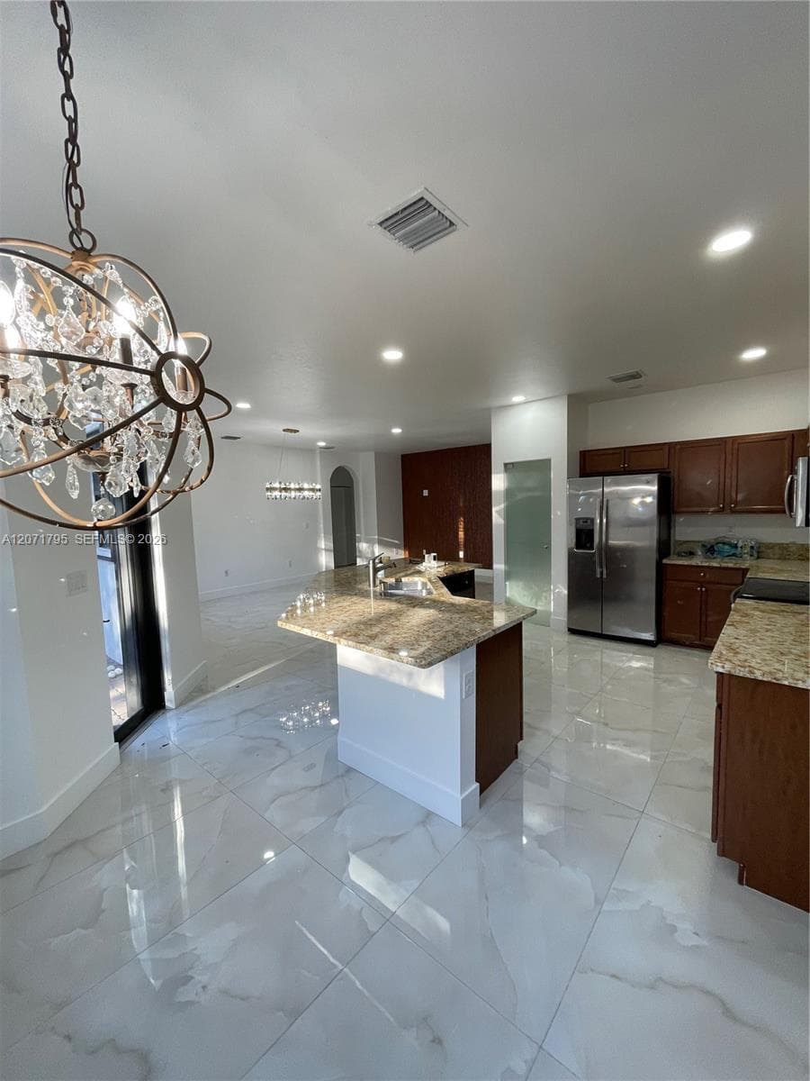 Kitchen featuring stone-finish countertops, dark wood cabinetry, large-format tile flooring, a center island with an integrated sink, and an ornamental metal and crystal chandelier