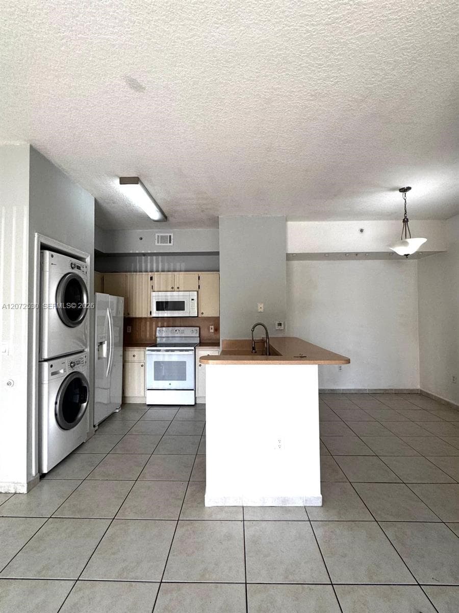 Kitchen area featuring light-toned wood cabinetry, an integrated island with a sink, white appliances, and ceramic tile flooring