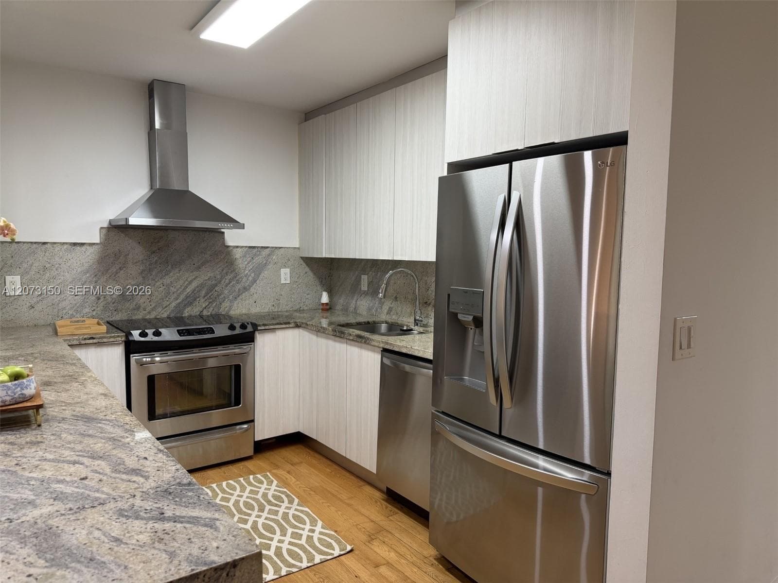 Kitchen featuring flat-panel cabinetry, stone countertops with a matching full-height backsplash, wood-finish flooring, and stainless steel appliances including a built-in range hood