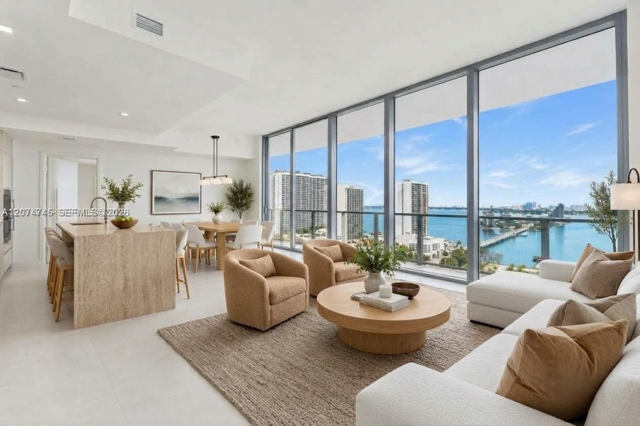 Open-concept living space featuring floor-to-ceiling windows, waterfront balcony access, neutral stone-finish kitchen island, and recessed lighting