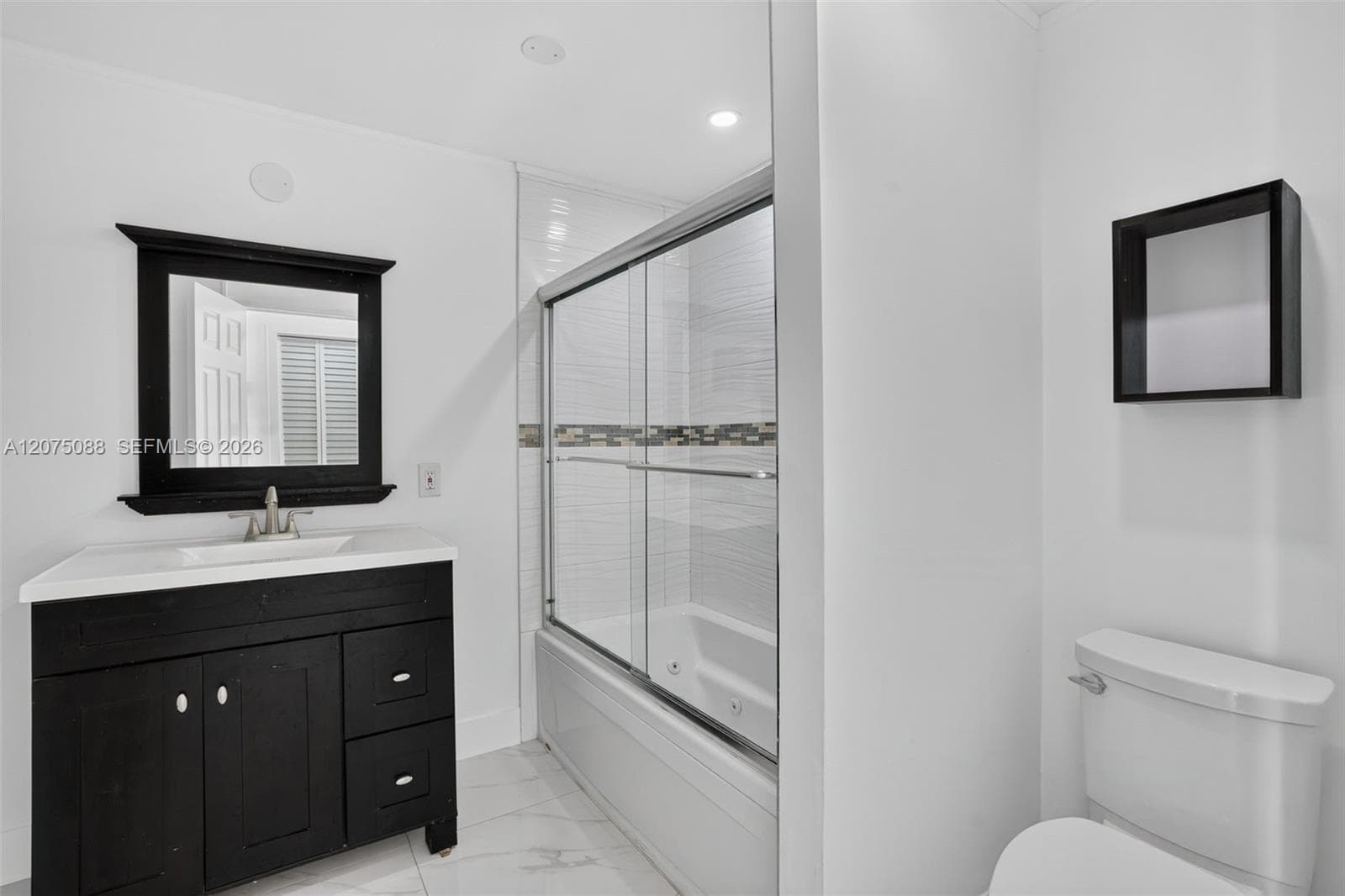 Bathroom featuring a dark-finish vanity with an integrated basin, a black-framed wall mirror, a glass-enclosed tub with tiled surround, and a wall-mounted shelving unit