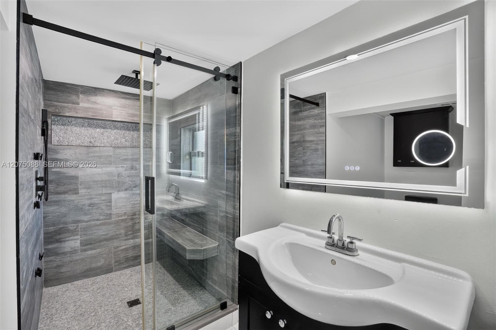 Walk-in shower featuring grey stone-finish tile, glass sliding enclosure, built-in bench, and rainfall showerhead, paired with a modern vanity and backlit wall mirror