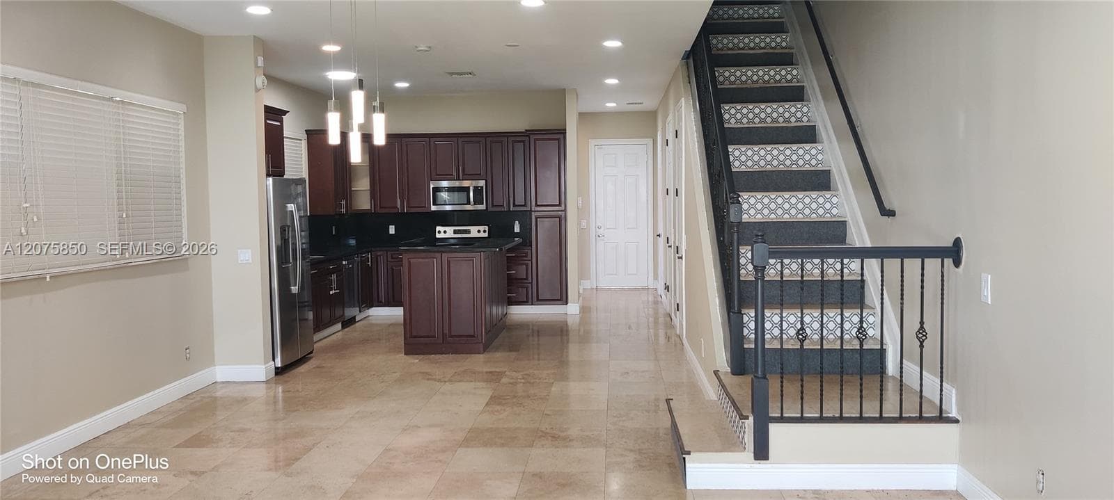 Kitchen featuring dark wood cabinetry, a central island, and stainless steel appliances, set against stone-finish flooring and a staircase with metal railing