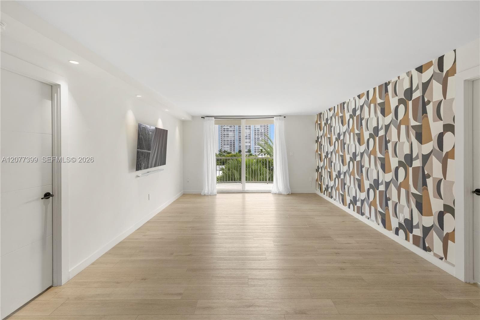 Open-concept interior featuring wood-finish flooring, recessed lighting, a geometric accent wall, and floor-to-ceiling glass doors opening to an exterior balcony