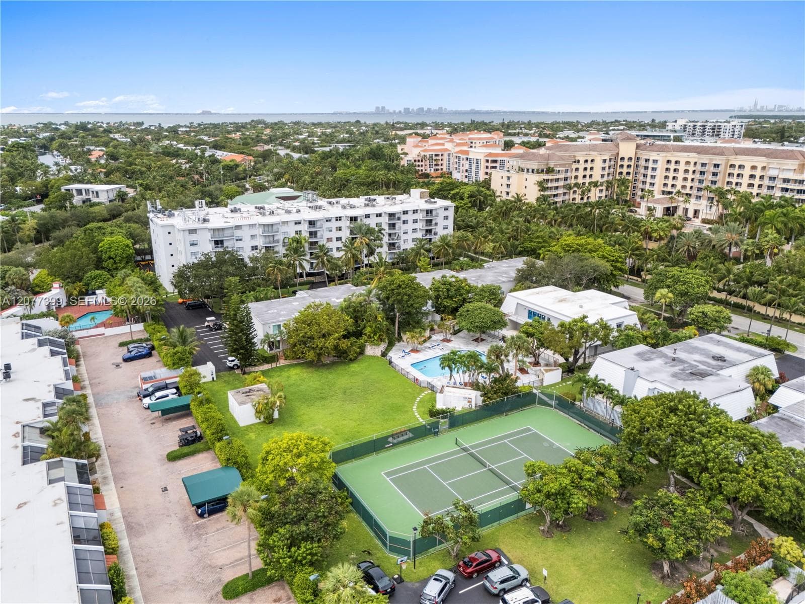 Aerial perspective of community grounds featuring a tennis court, swimming pool, manicured lawn, and mature canopy trees