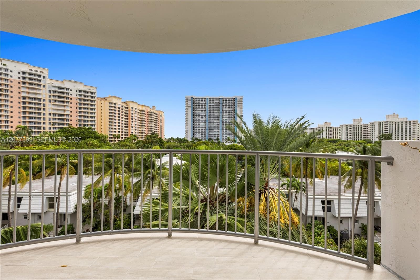Covered balcony with neutral tile flooring and metal railings, featuring mature palms and surrounding mid-rise residential architecture