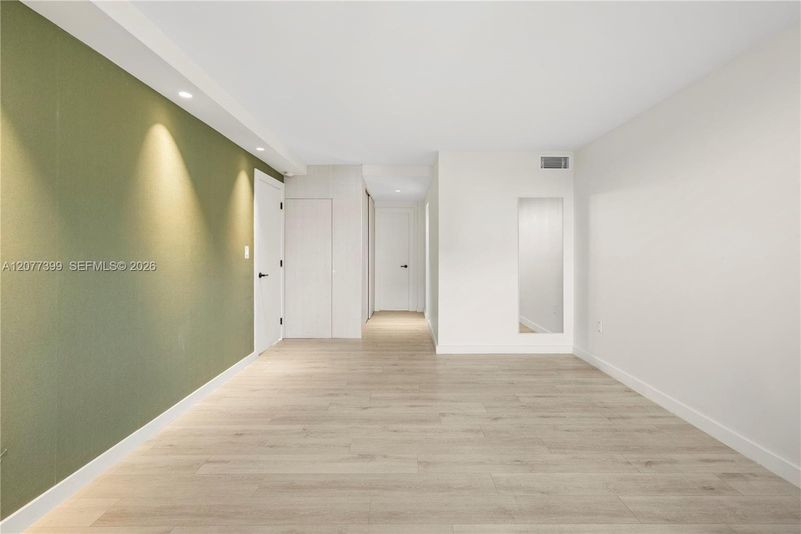 Interior room featuring light wood-finish flooring, recessed ceiling lighting, a prominent sage green accent wall, and a full-length wall-recessed mirror