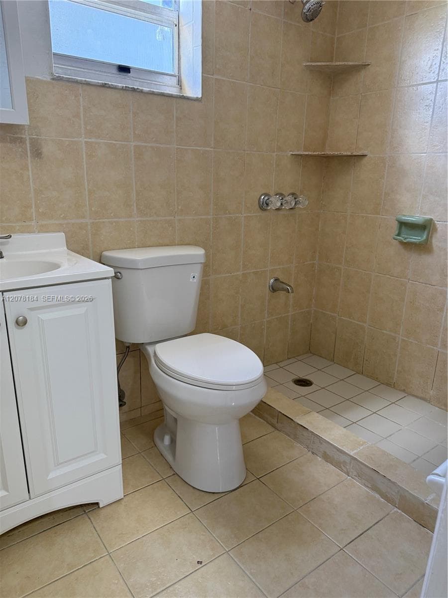 Bathroom featuring a white vanity cabinet, ceramic tile flooring, a step-in tiled shower with corner shelving, and a wall-mounted window