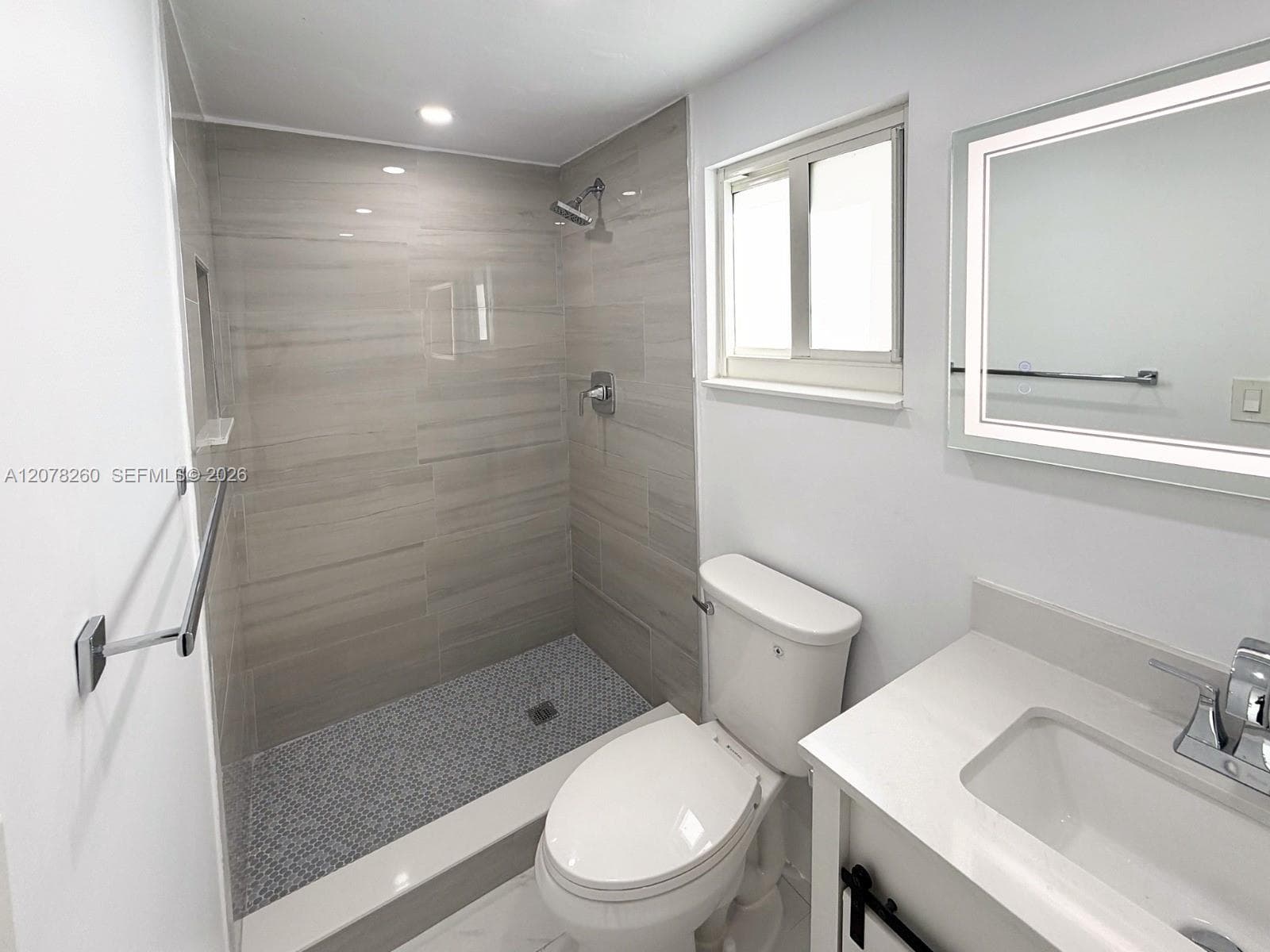 Walk-in shower featuring light-toned large-format tile surround and mosaic tile flooring, vanity with integrated basin and quartz countertop, illuminated mirror, and polished chrome fixtures