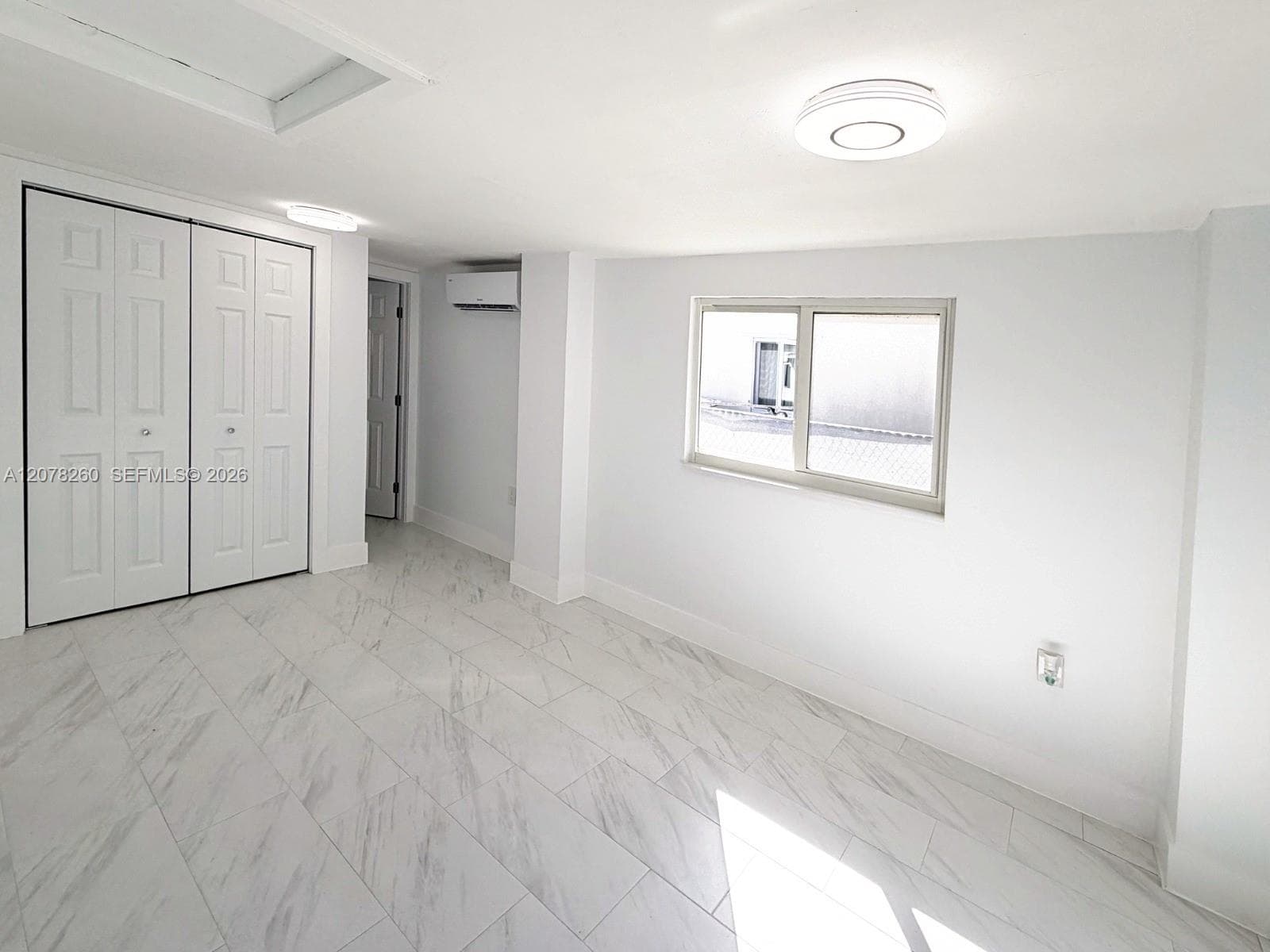 Neutral-toned space featuring large-format tile flooring, dual-panel closet doors, a wall-mounted ductless climate control unit, and flush-mount ceiling lighting