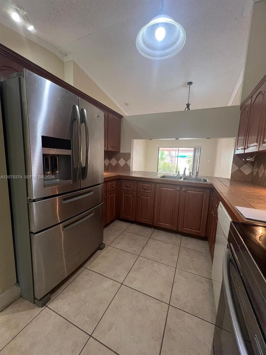 Kitchen featuring stainless steel refrigerator, wood cabinetry, tile countertops with diamond-patterned backsplash, and ceramic floor tile