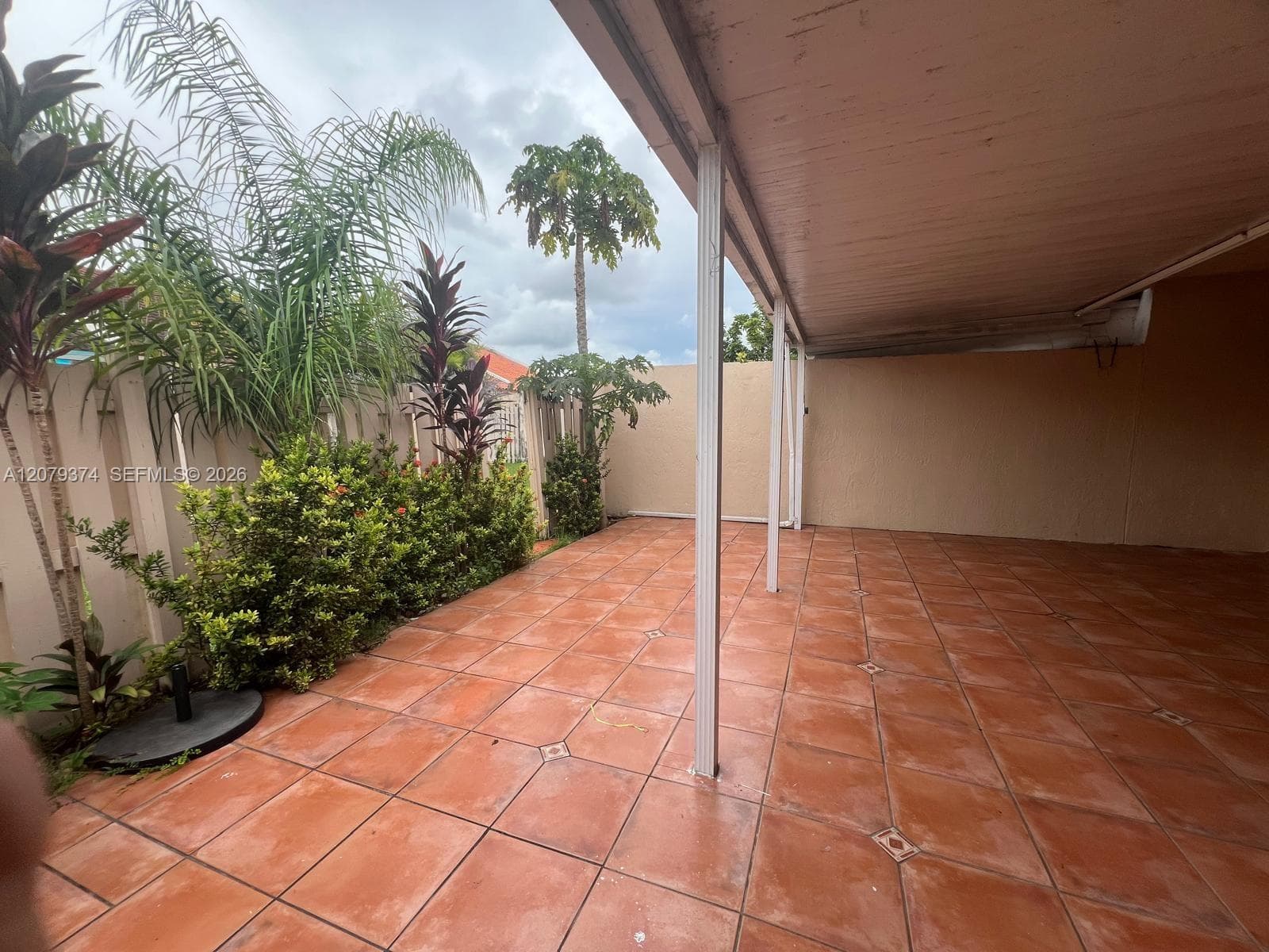 Covered patio featuring terracotta-style tile flooring, metal support columns, and perimeter privacy wall with mature tropical landscaping