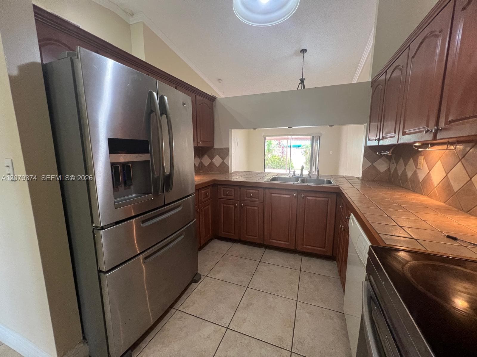 Kitchen featuring wood cabinetry, tile countertops, tile backsplash, stainless steel refrigerator, and tile flooring