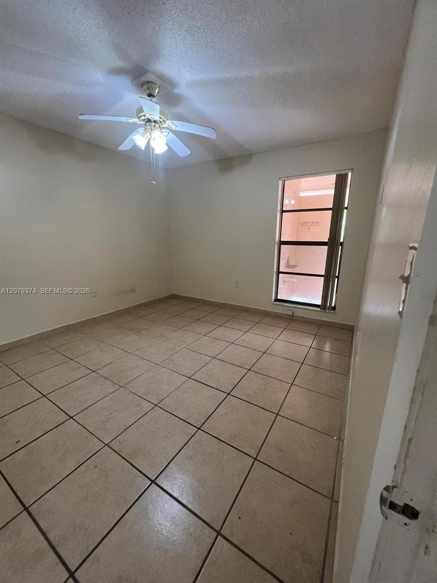 Room featuring neutral-toned tile flooring, a multi-blade ceiling fan with integrated lighting, and a single-pane window with dark metal framing