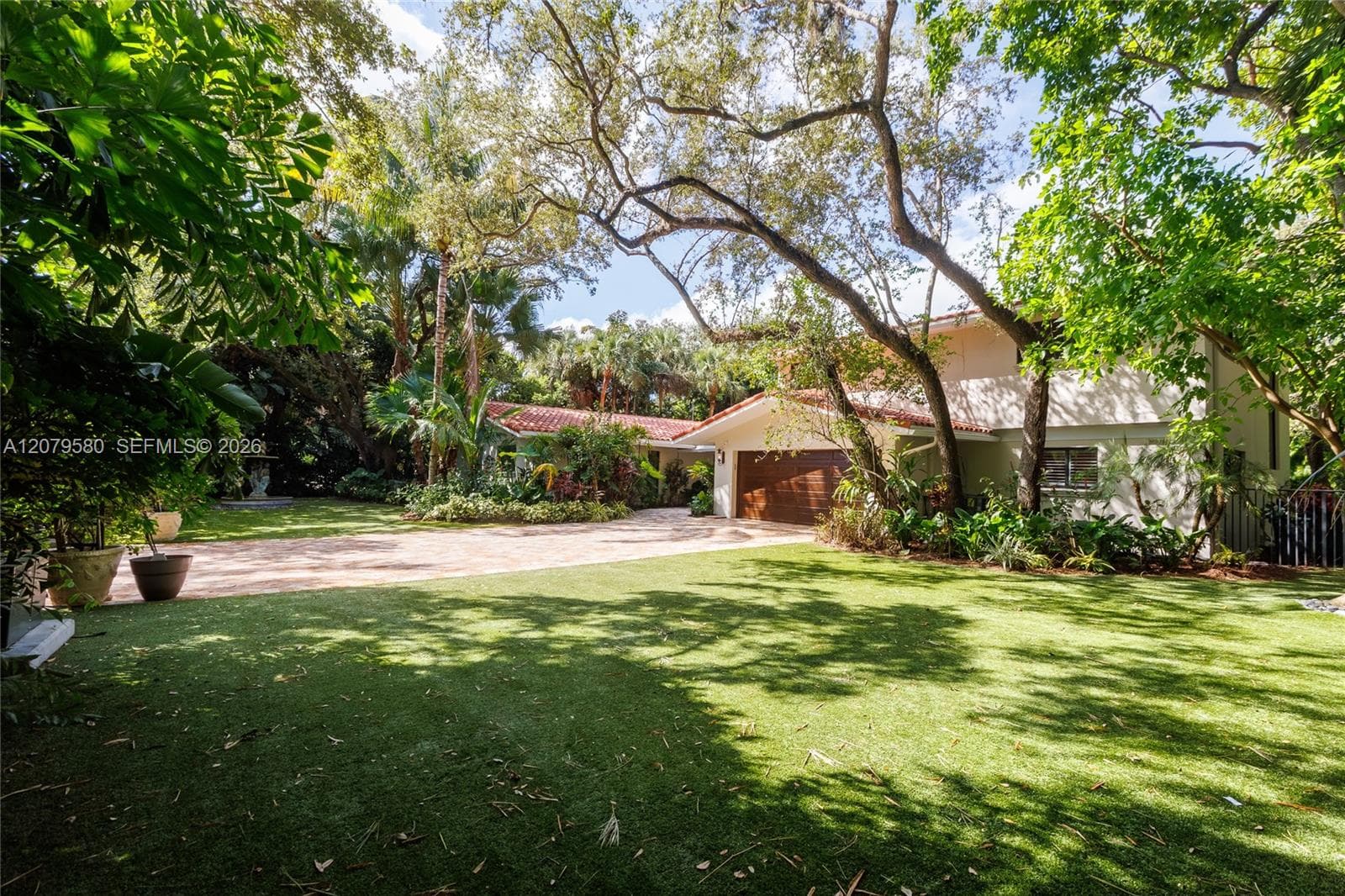 Exterior featuring a low-pitch tile roof, expansive front lawn, paved driveway, multi-car garage with horizontal wood-finish door, and established mature trees