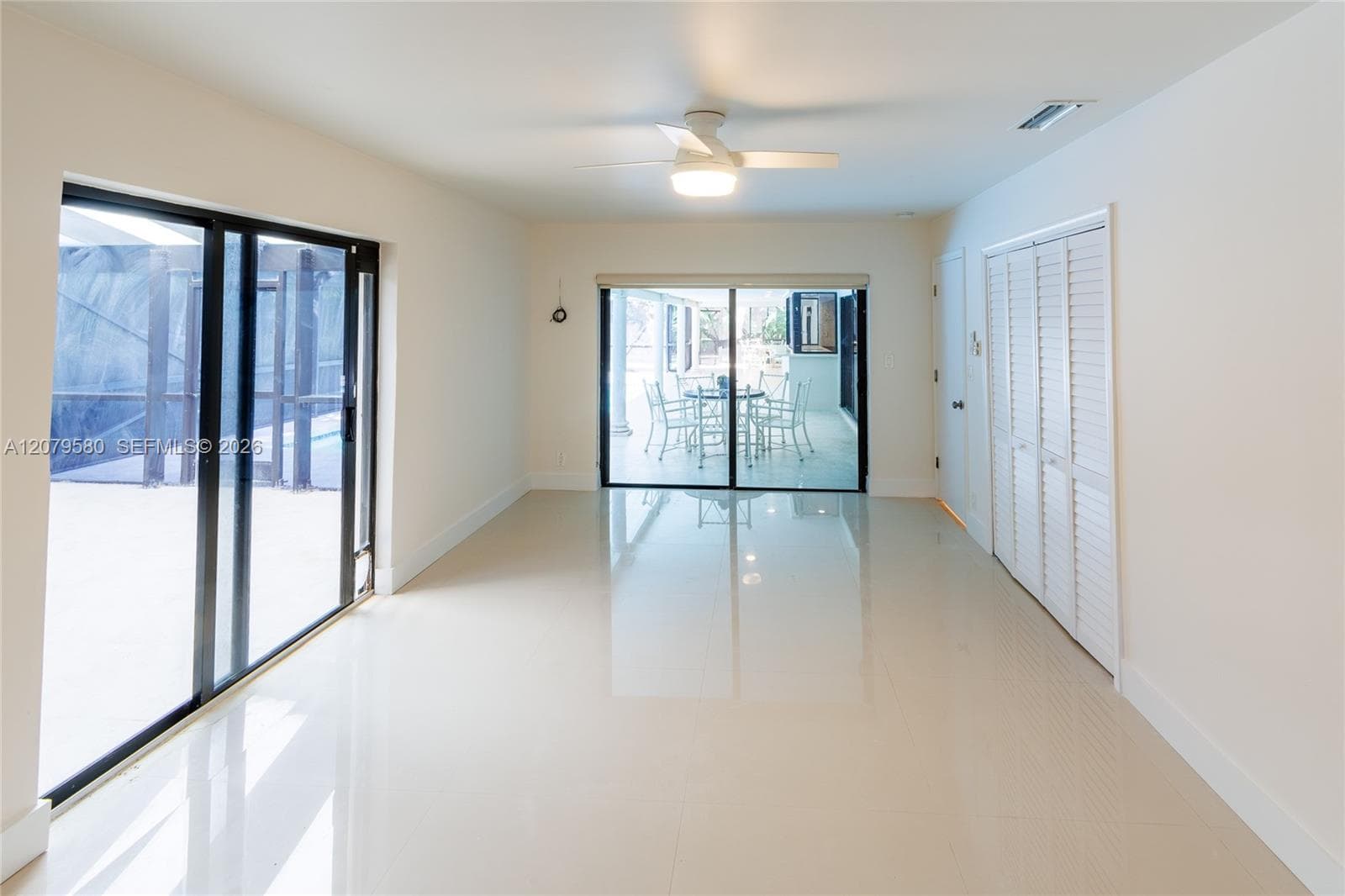 Interior space featuring light-toned tile flooring, dual sets of sliding glass doors with black frames, white louvered closet doors, and a central ceiling fan with integrated lighting