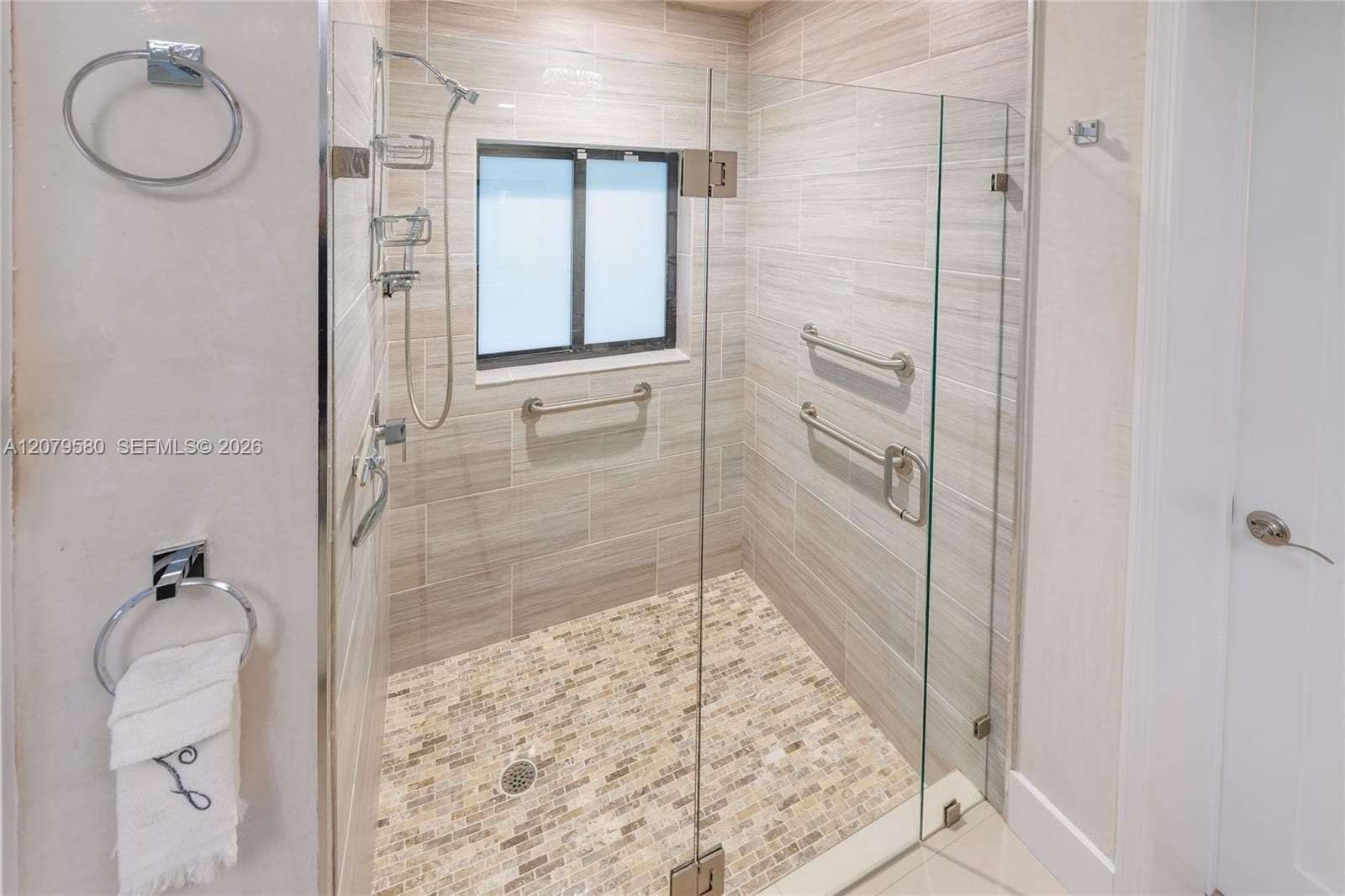 Walk-in shower featuring a frameless glass door, horizontal wall tiling, stone-mosaic floor tile, built-in metal grab bars, and a hand-held showerhead